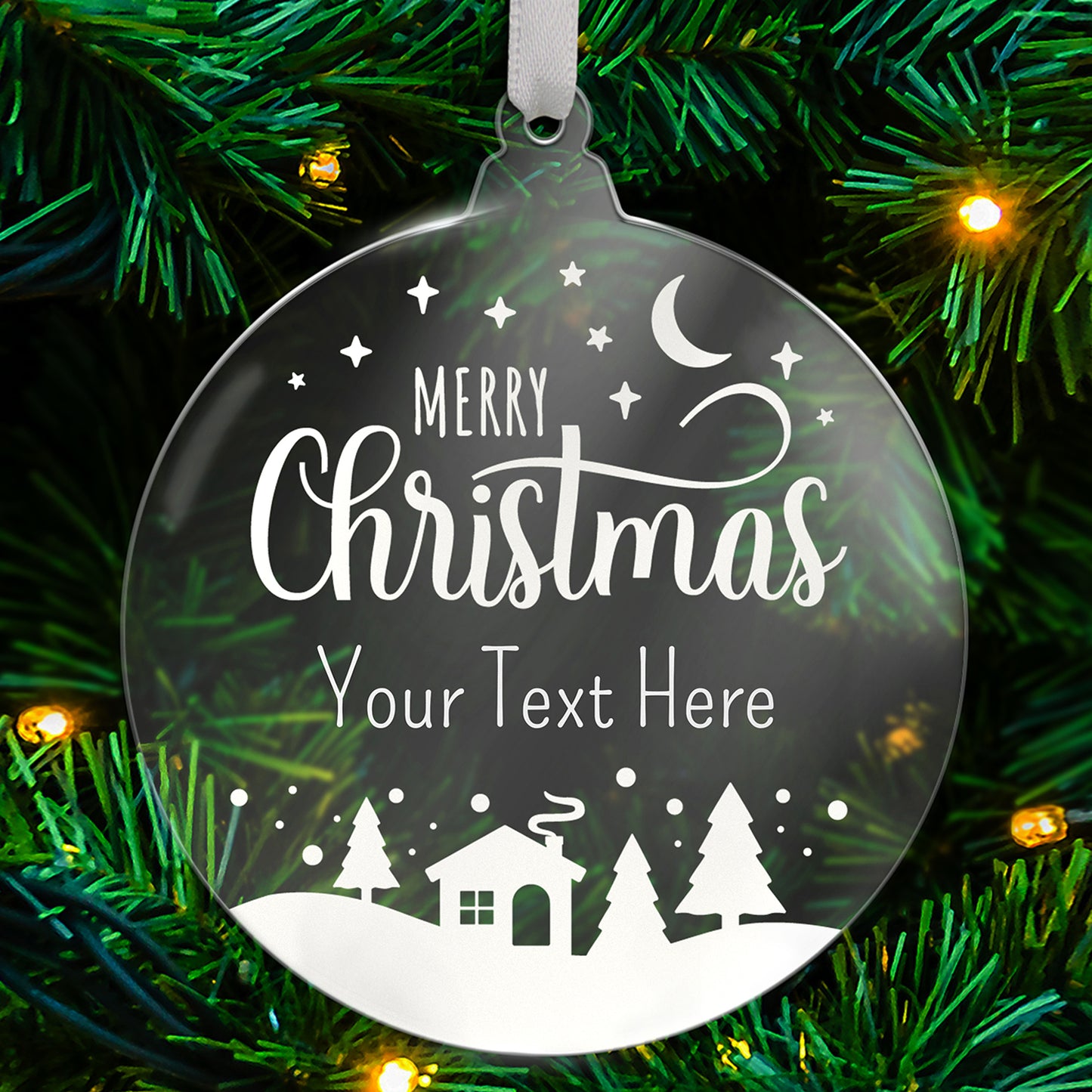 A round, clear acrylic Christmas tree ornament hanging from a white ribbon. The ornament features a festive winter design with the words 'Merry Christmas' in elegant white script, followed by placeholder text 'Your Text Here'. Below, there's a silhouette of a snowy landscape with pine trees, a house with smoke rising from the chimney, and a crescent moon with stars in the sky. The ornament is displayed against a green Christmas tree with lights.