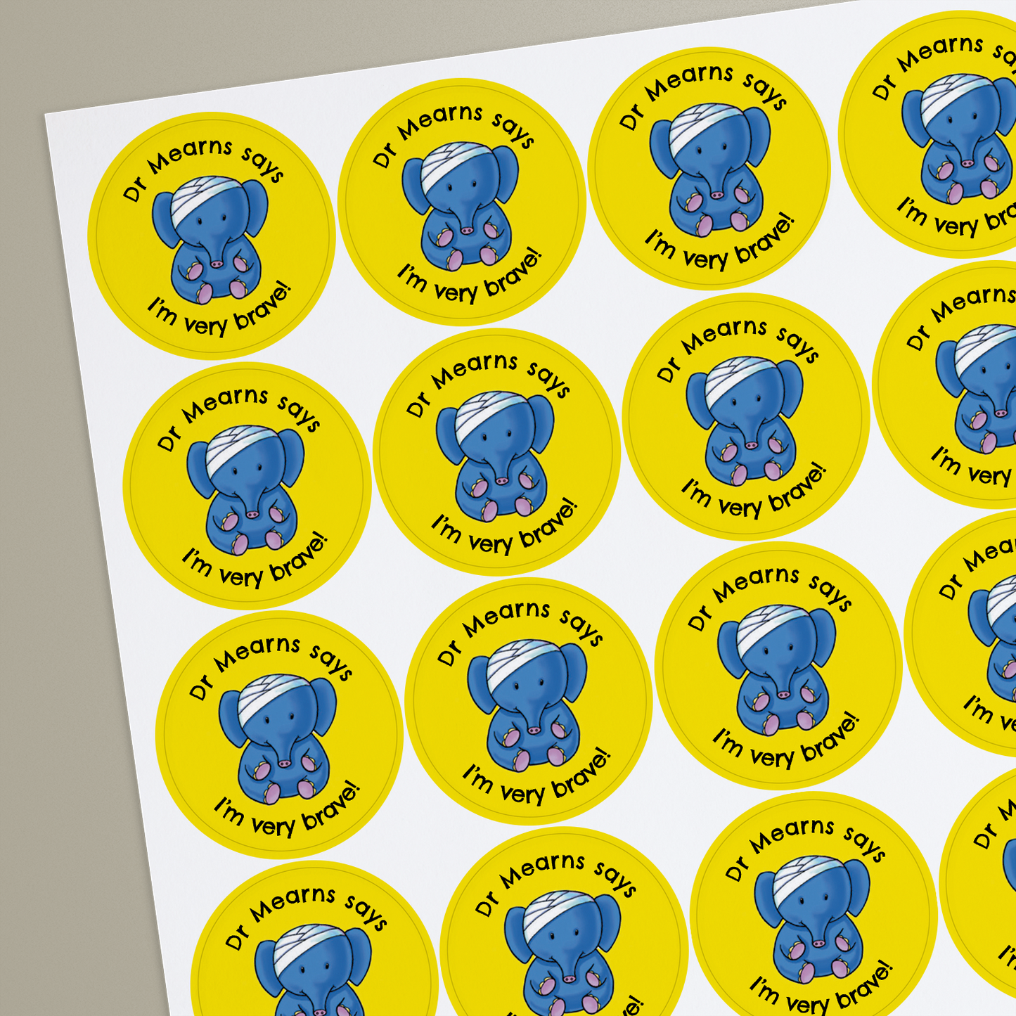 Sheet of round yellow stickers featuring an elephant with a bandage on it's head in the centre. Each sticker has placeholder text reading 'Dr Mearns says I'm very Brave!' curved above and below the illustration.
