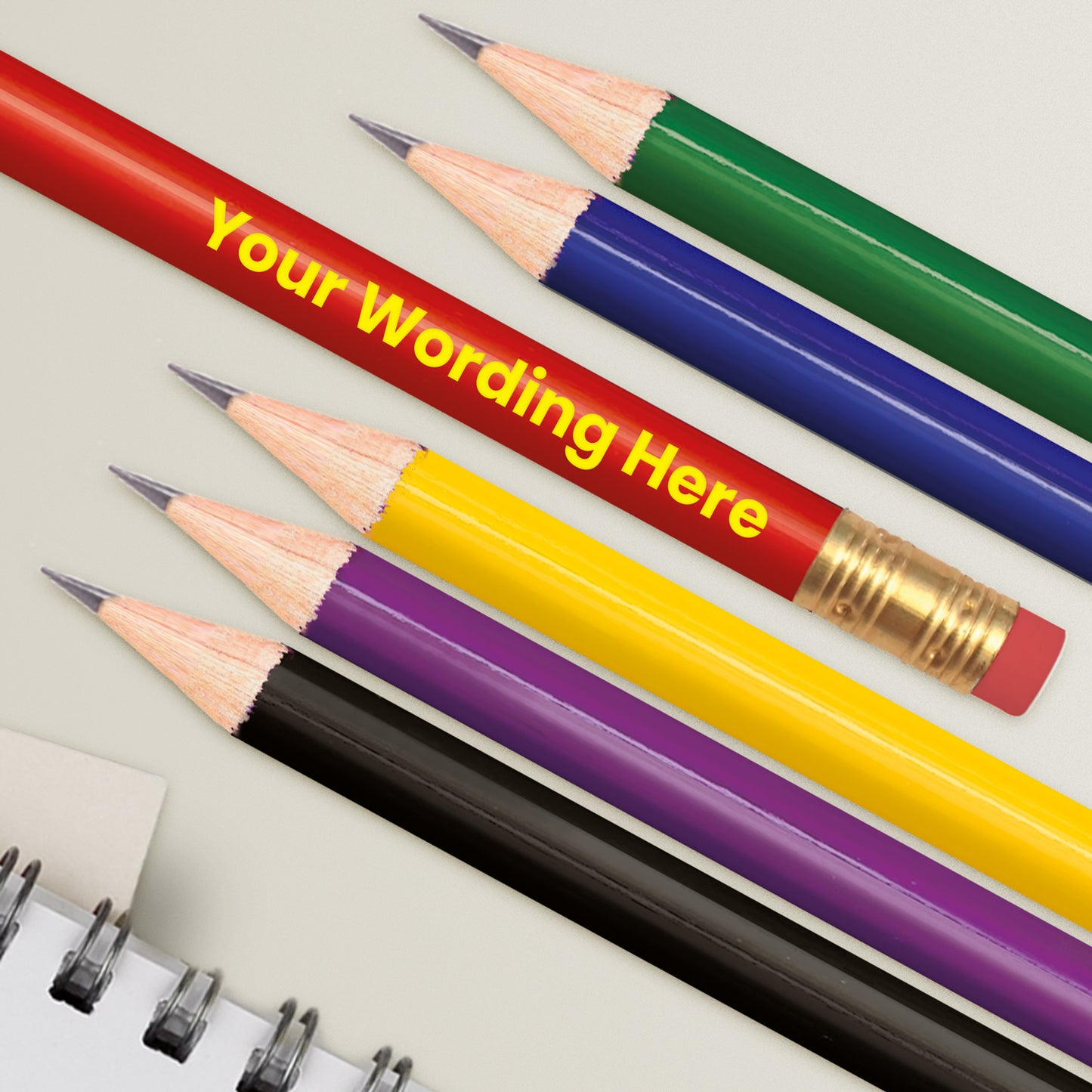 A set of multi-colored personalised HB pencils with custom text 'Your Word Here' printed on each, positioned alongside a eraser and a notepad.