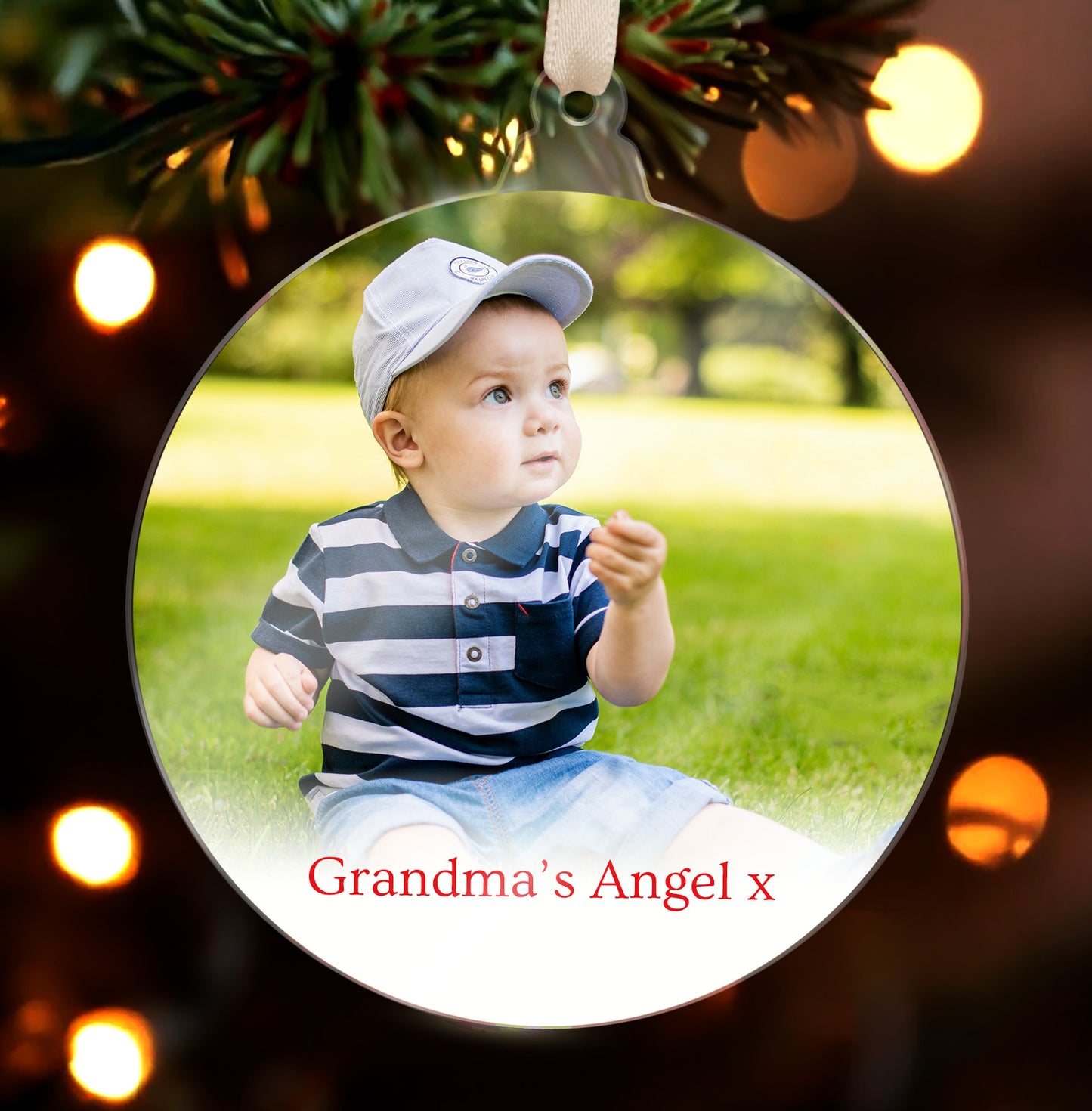Personalised acrylic Christmas tree ornament featuring an uploaded photograph of a child with text saying Grandma's Angel x, with festive evergreen branches and lights in the background.
