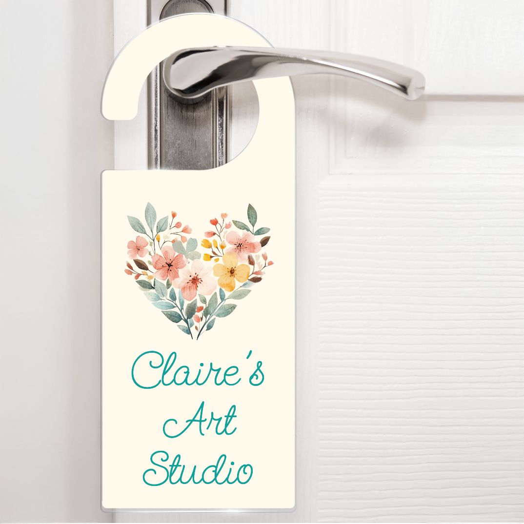 A cream door hanger hanging on a silver handle, featuring a bouquet of flowers shaped like a heart above the text 'Claire's Art Studio' in scrolling teal letters. The design is artistic and sophisticated