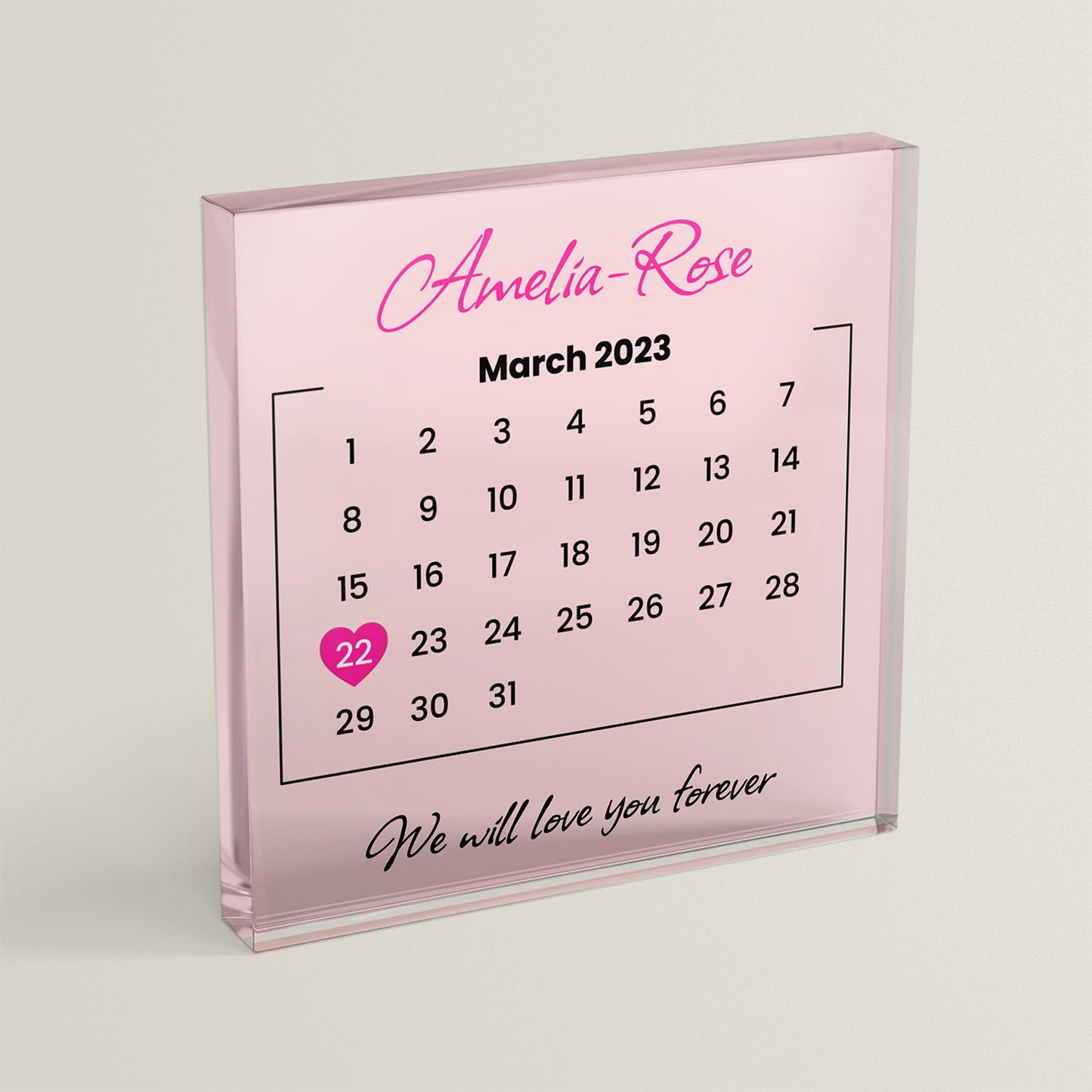 A pale pink acrylic block on a cream background, showing example text "Amelia-Rose" at the top, a calendar with the date 22nd March 2023 highlighted with a heart and example text "We will love you forever" at the bottom.