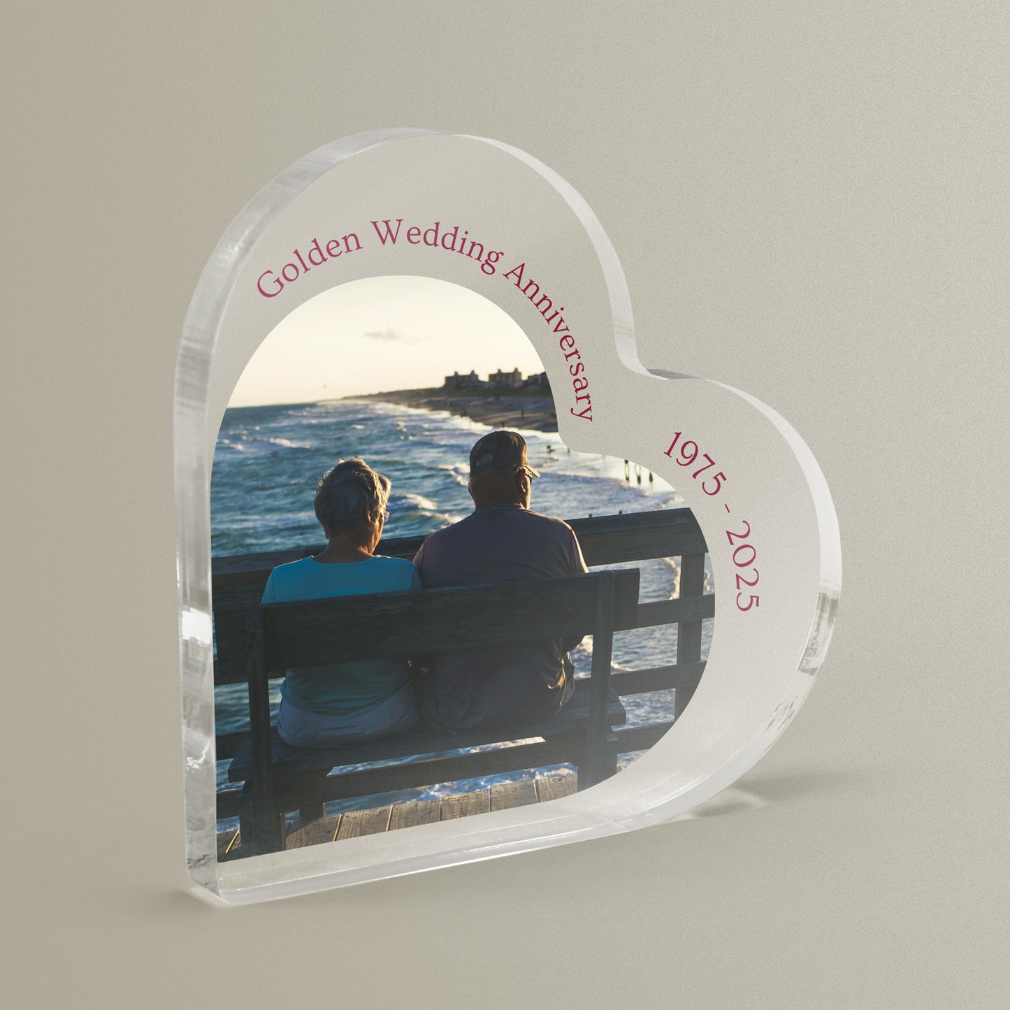 Heart-shaped acrylic plaque with a photo of two people sitting on a bench by the sea with personalised wording. Block is shown against a grey background.