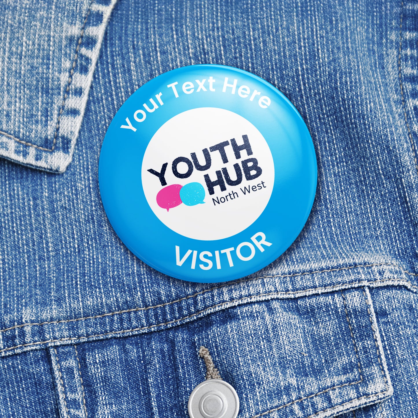 A blue circular business badge with a logo and the text 'YOUTH HUB North West Visitor' displayed on it, pinned to a denim fabric.