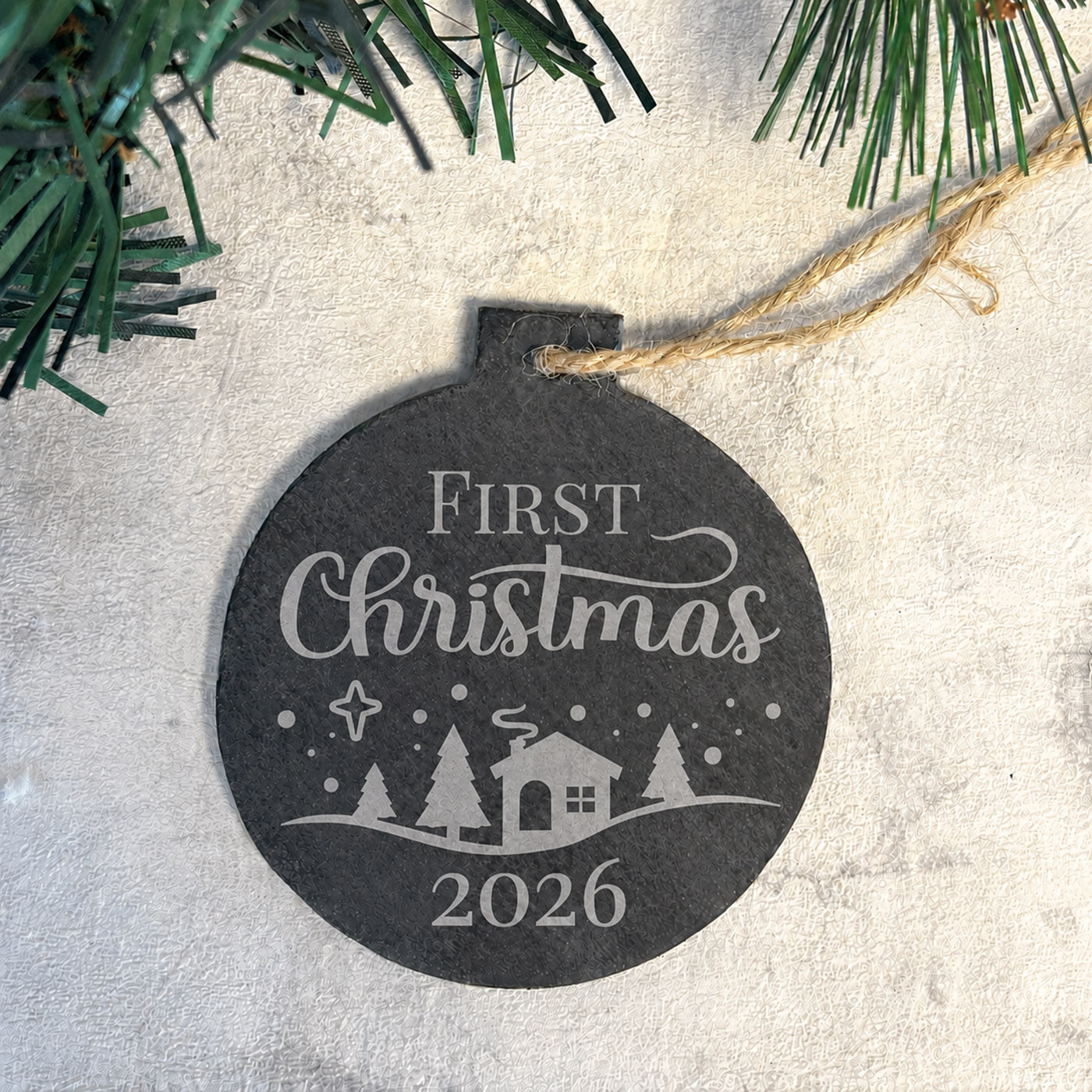 Round Christmas ornament with 'First Christmas 2026' text and house design, hanging on a branch.