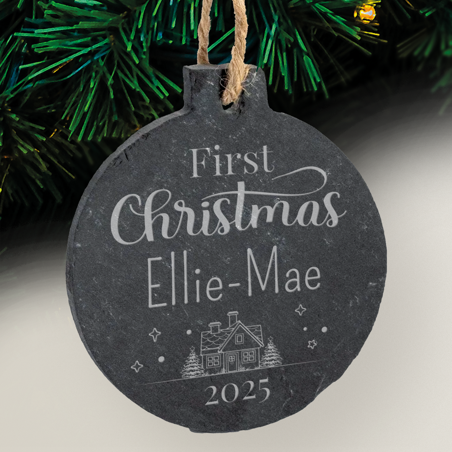 Round slate ornament with 'First Christmas Ellie-Mae 2025' text, house illustration, and tree in the background.