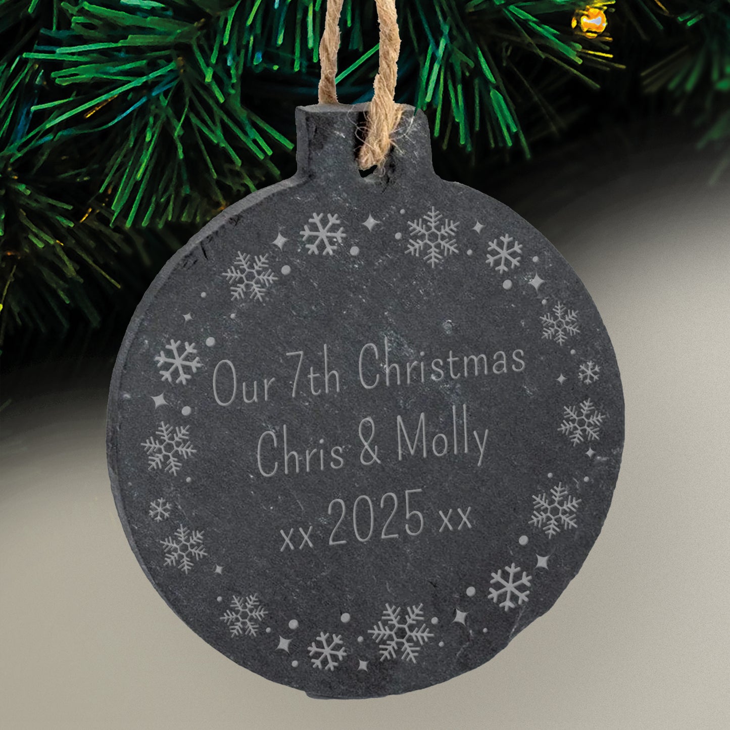 Slate Christmas bauble ornament with a wreath design etched around the edge and 'Our 7th Christmas Chris & Molly xx 2025 xx' text within it, hanging against a festive background.
