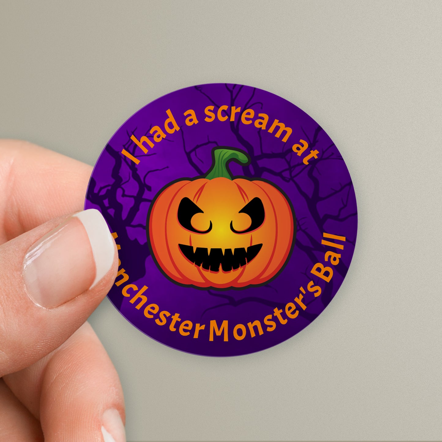Hand holding a round, purple sticker with personalised text above and below a carved pumpkin.