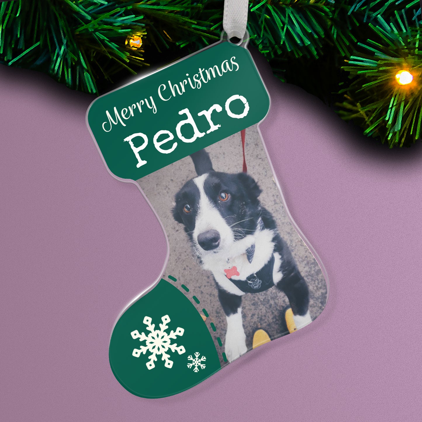 Stocking-shaped acrylic Christmas tree ornament with a deep green design, white snowflake decorations, and the message 'Merry Christmas Pedro' at the top, featuring a photo of black and white dog, hanging on a tree with fairy lights.