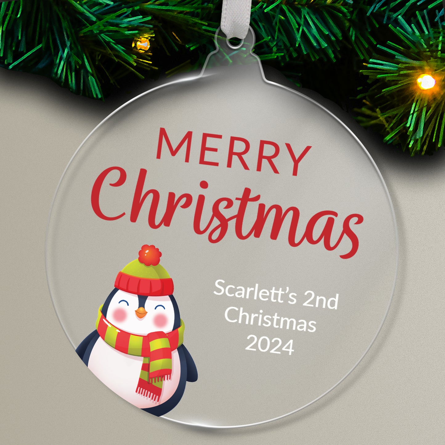 Round acrylic Christmas tree ornament featuring a festive penguin wearing a hat and scarf, with the message 'Merry Christmas' in red and space for custom text below, hanging on a tree with green needles and lights.