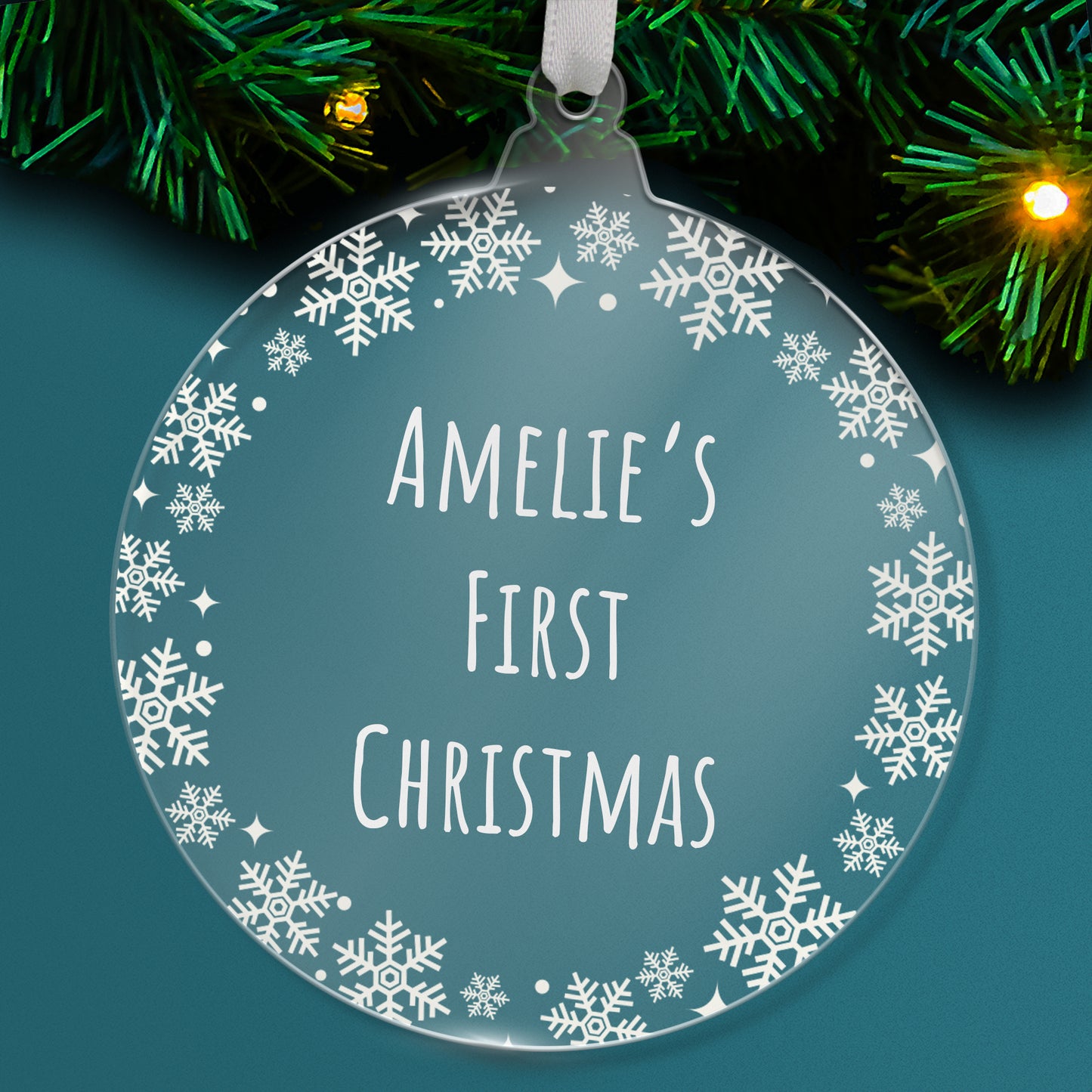 Round acrylic clear Christmas tree ornament with decorative white snowflakes around the border, featuring the words 'Amelie's First Christmas' in a tall, narrow font, hanging on a tree with green branches and fairy lights.