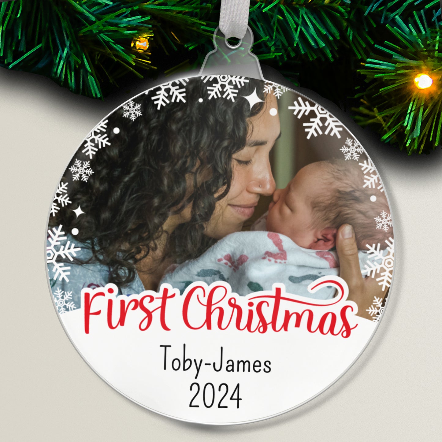 A personalised photo upload first Christmas bauble with a photo of a mother holding her baby, and text 'Toby James' on it, surrounded by snowflakes.
