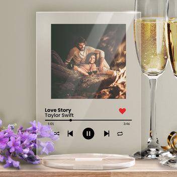 A personalised acrylic music plaque styled like a digital music player. It features a photo of a couple on the beach at the top, followed by song details lines for the song title and artist. Music player controls, a progress bar, and a red heart icon appear below, mimicking a streaming app interface. The plaque stands upright on a clear circular base.