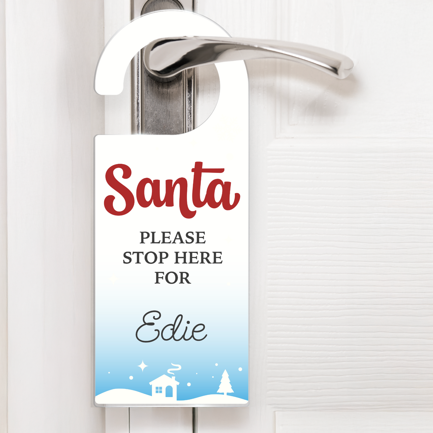 A festive snow scene door hanger with white text that reads 'Santa PLEASE STOP HERE FOR Edie'. The design includes snowflakes, stars, and a snowy village scene with a house and trees at the bottom. The hanger is displayed on a silver door handle, ready for Christmas Eve.
