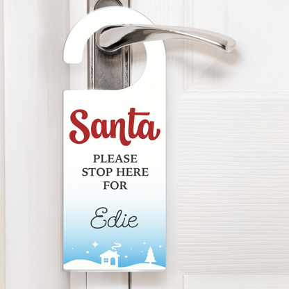 A festive snow scene door hanger with white text that reads 'Santa PLEASE STOP HERE FOR Edie'. The design includes snowflakes, stars, and a snowy village scene with a house and trees at the bottom. The hanger is displayed on a silver door handle, ready for Christmas Eve.
