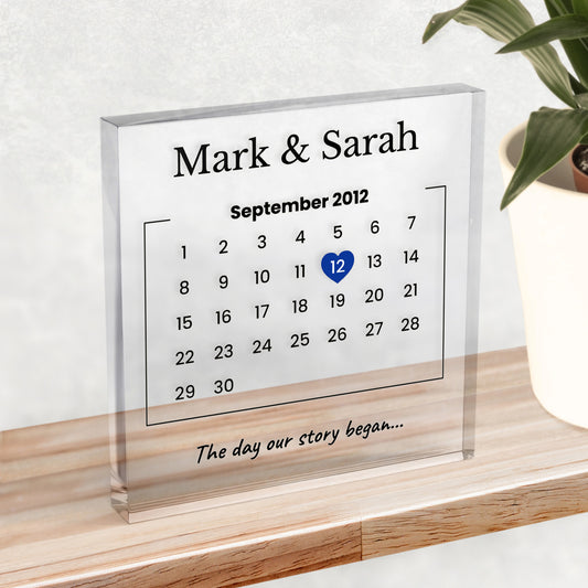 A clear acrylic block on a shelf, showing names at the top, a calendar with the date 12th September 2012 highlighted with a heart and example text "The day our story began..."
