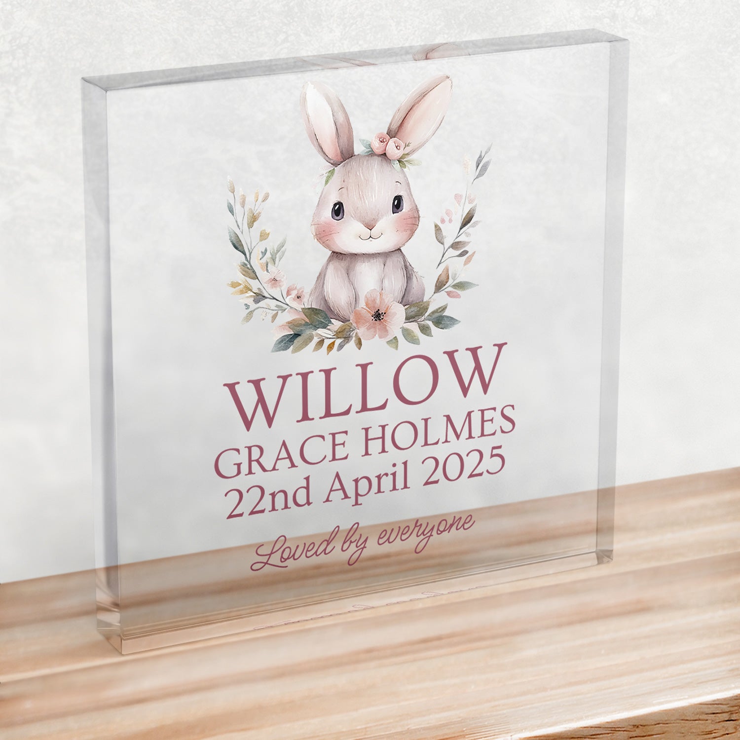 A clear plastic square block on a wooden shelf, showing an illustrated rabbit at the top, wording below reads, “WILLOW GRACE HOLMES, 22nd April 2025, Loved by everyone”