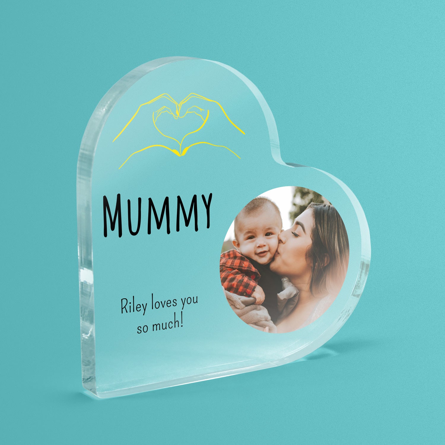 Heart-shaped acrylic block with a photo of a woman kissing a baby, hands making a heart and text on a teal background.