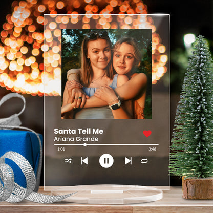 A personalised acrylic music plaque styled like a digital music player. It features a photo of two women of at the top, followed by song details lines for the song title and artist. Music player controls, a progress bar, and a red heart icon appear below, mimicking a streaming app interface. The plaque stands upright on a clear circular base.