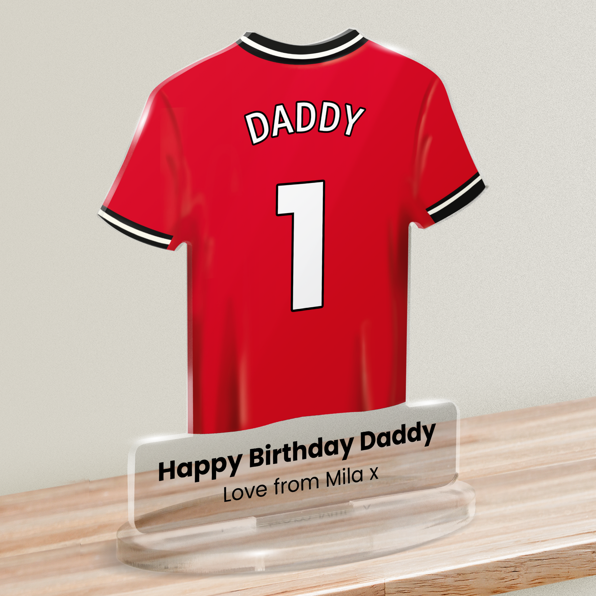 Acrylic t-shirt plaque with a red sports shirt design with 'DADDY' and number '1' on the shirt and 'Happy Birthday Daddy' 'Love from Mila x' underneath. Plaque is shown on a wooden shelf against a white background.