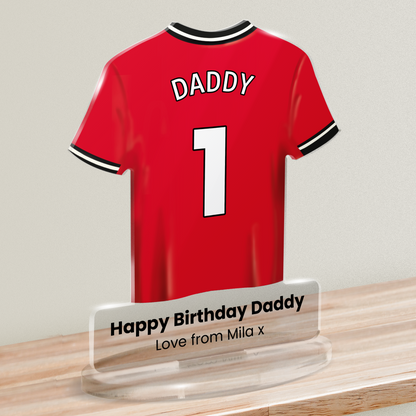 Acrylic t-shirt plaque with a red sports shirt design with 'DADDY' and number '1' on the shirt and 'Happy Birthday Daddy' 'Love from Mila x' underneath. Plaque is shown on a wooden shelf against a white background.