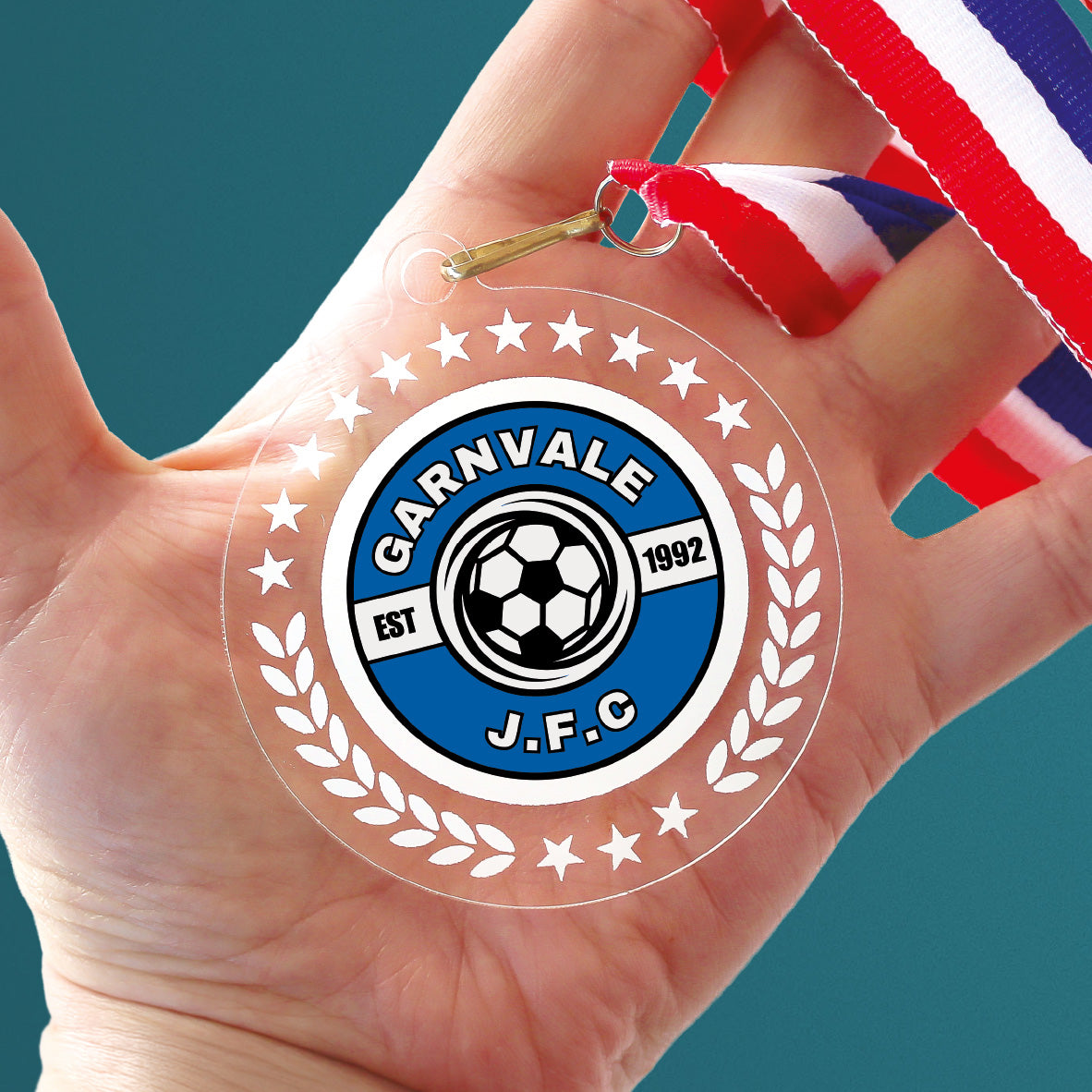 Acrylic personalised medal with stripy ribbon showing clear background with wreath design and 'Garnvale JFC' logo. held in a hand.