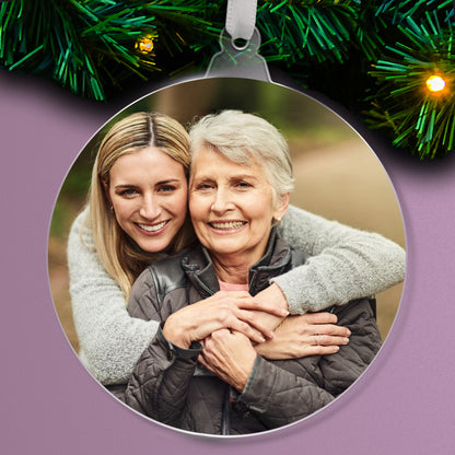 Personalised acrylic Christmas tree ornament featuring an uploaded photograph of a mother and daughter, with festive evergreen branches and lights in the background.
