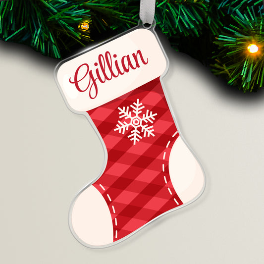 Clear acrylic Christmas stocking-shaped tree ornament decorated with a white snowflake on a red checked background featuring a space at the top for a personalised name in red script, hanging on a Christmas tree with lights.
