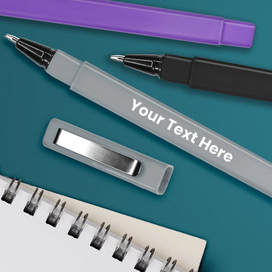 Personalised grey ballpoint pen with white text, shown with black and purple pens and a notebook on a teal background.